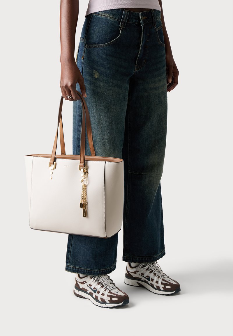 Un sac fourre-tout blanc avec des poignées en cuir marron, présentant des éléments en or, un charme en forme de pendentif et un design structuré. Porté avec un jean large bleu et des baskets.