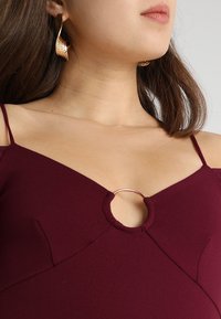 Vínová šaty s ramínky na spaghetti, s výřezem ve tvaru klíče zdobeným kovovým kroužkem. Hladký texturovaný materiál, úzký střih.