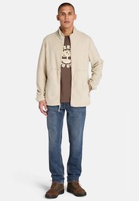 Chaqueta de forro polar beige con cuello alto, cierre de cremallera y bolsillos laterales, usada sobre una camiseta gráfica marrón y combinada con vaqueros azules y zapatos marrones.