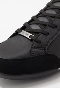 Sneaker nero con una combinazione di pelle liscia e texture scamosciata, lacci neri piatti e un accento di logo metallico argento.
