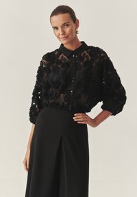 Camicia nera trasparente decorata con applicazioni floreali testurizzate, dotata di colletto, chiusura con bottoni e maniche ampie, abbinata a una gonna nera.