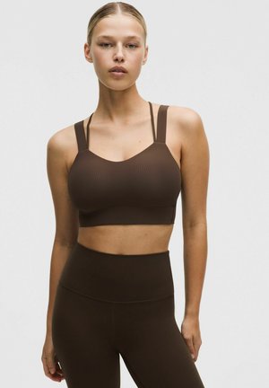 Brązowy sportowy biustonosz z prążkowanego materiału, z szerokimi ramiączkami, dopasowanym krojem i dekoltem w kształcie litery U, zestawiony z brązowymi legginsami z wysokim stanem. Gładka faktura.