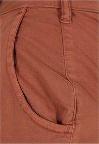 Gros plan d'une poche de pantalon en tissu couleur rouille avec coutures visibles et détails des passants de ceinture.