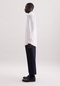 Camicia bianca a maniche lunghe con polsini abbottonati, abbinata a pantaloni blu navy e scarpe eleganti nere, presentata in profilo laterale su uno sfondo chiaro.