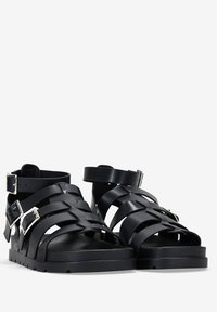 Sandalias negras con un diseño de tiras trenzadas, hechas de material sintético, con hebilla de metal plateado y suela plana de goma.