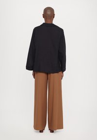Giacca oversized nera in lino con maniche lunghe, colletto a scuto e vestibilità ampia, abbinata a pantaloni marroni a gamba larga.