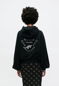 Μαύρ ο φούτερ με κουκούλα, με λευκό γραφικό κείμενο και αστέρια στην πλάτη, σε συνδυασμό με μαύρη φούστα με σχέδιο διαμαντιών.