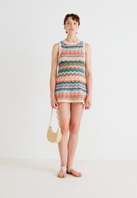Multifarget strikket ermeløs kjole med zigzag-mønster, med bølgede kanter. Kombinert med en rund gullhåndveske og beige sandaler.