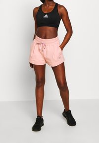 Černý sportovní crop top s bílým logem, spárovaný se světle růžovými šortkami s elastickým pasem a předními kapsami. Černé tenisky.