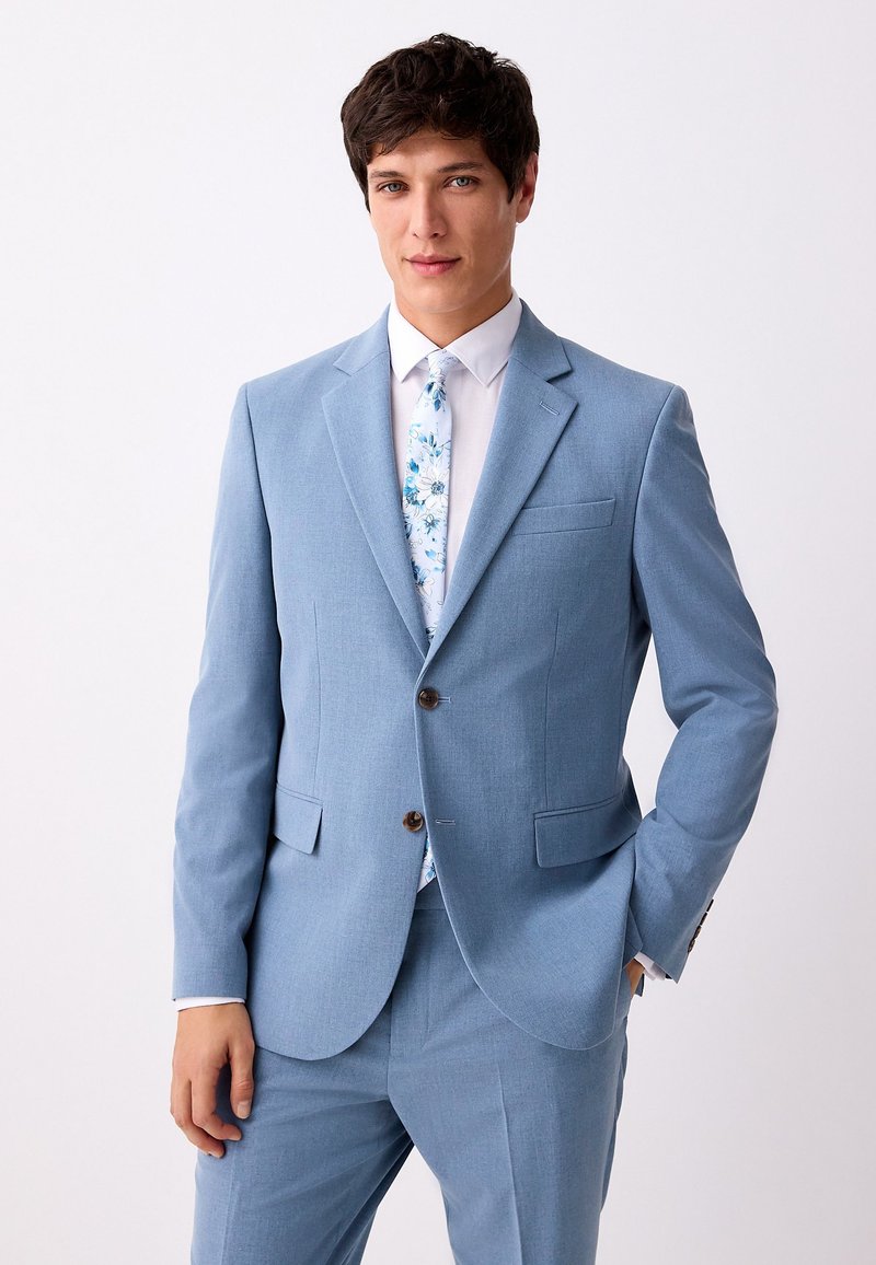 Abito azzurro chiaro con rever a incavo, due bottoni e tasche con patta; abbinato a una camicia bianca e una cravatta floreale con motivi blu e bianchi.