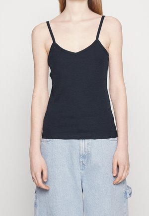 Vrouw draagt een zwart mouwloos V-hals tanktop gecombineerd met lichtblauwe loszittende spijkerbroek tegen een effen achtergrond.