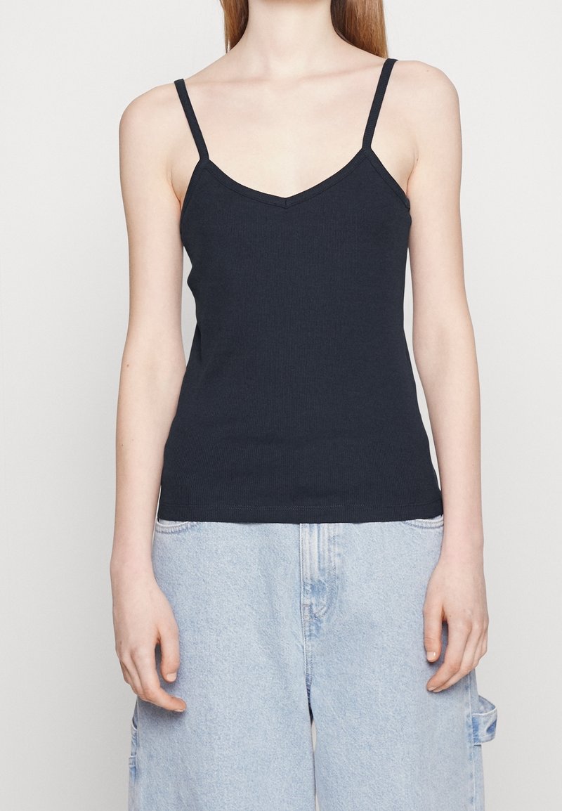Femme portant un débardeur noir sans manches à col en V associé à un jean en denim bleu clair ample sur un fond uni.