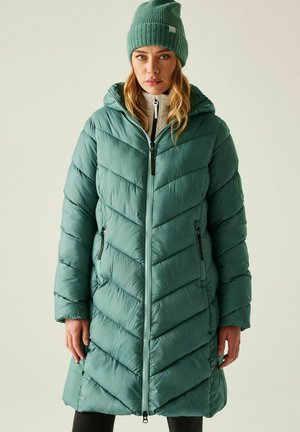 Vrouw draagt een teal gekleurde donsjas met diagonale stiksels en capuchon, bijpassende teal gebreide muts, lichtgekleurde zip-up trui, staand tegen een effen achtergrond.