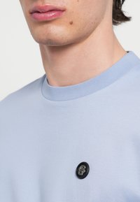 Svetlomodré tričko s krátkym rukávom, okrúhlym výstrihom a textúrovanou tkaninou. Na hrudi je čierne logo v podobe gombíka pre dodanie detailu.