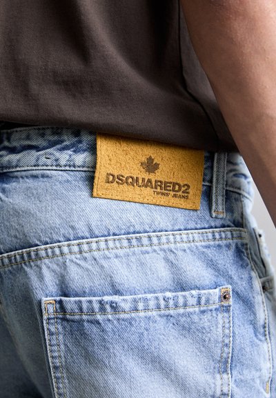 Svijetloplave traperice s žutim suedinim stražnjim pečatom na kojem se nalazi "DSQUARED2" i logo javora, prikazujući detalje šivanja i džepova.