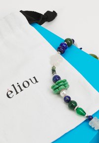 Perlenkette mit grünen, blauen und weißen Steinen, einschließlich Perlen, präsentiert auf einem weißen Stoffbeutel mit dem Markennamen "élou."