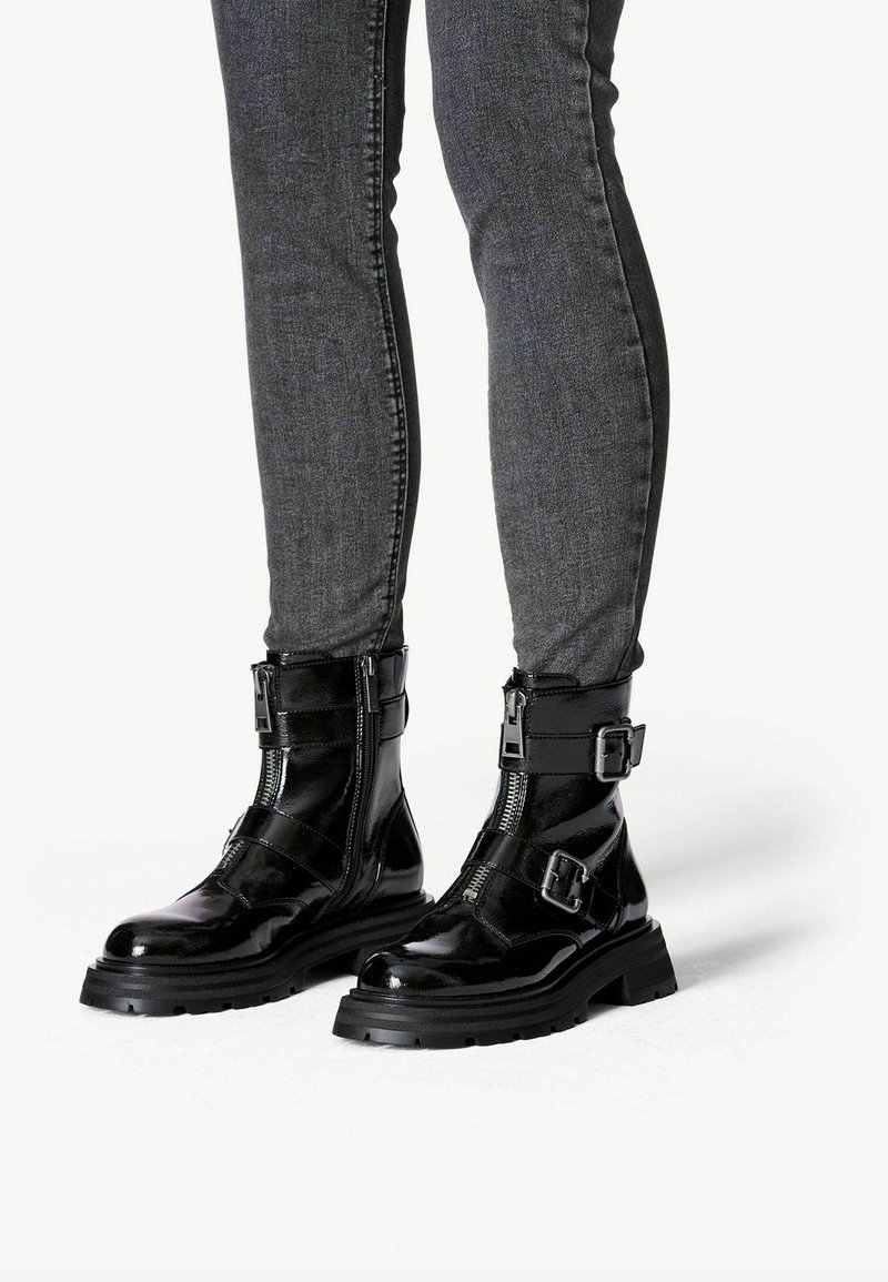 Bottes de combat noires brillantes avec des fermetures éclair latérales et des sangles à double boucle, portées avec un jean skinny gris foncé.