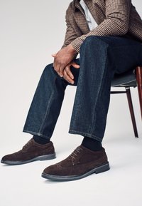 Chaussures habillées en daim marron avec des détails brogues, semelle noire plate, associées à un pantalon en denim foncé et une chemise à carreaux.