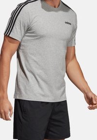 Maglietta atletica grigia a maniche corte, con scollatura a girocollo, logo Adidas nero e tre strisce nere sulle cuciture delle spalle.