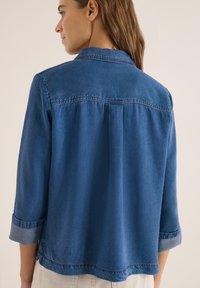 Femme aux cheveux bruns portant une chemise en denim bleue à manches longues avec les poignets retroussés, vue de dos sur un fond neutre.