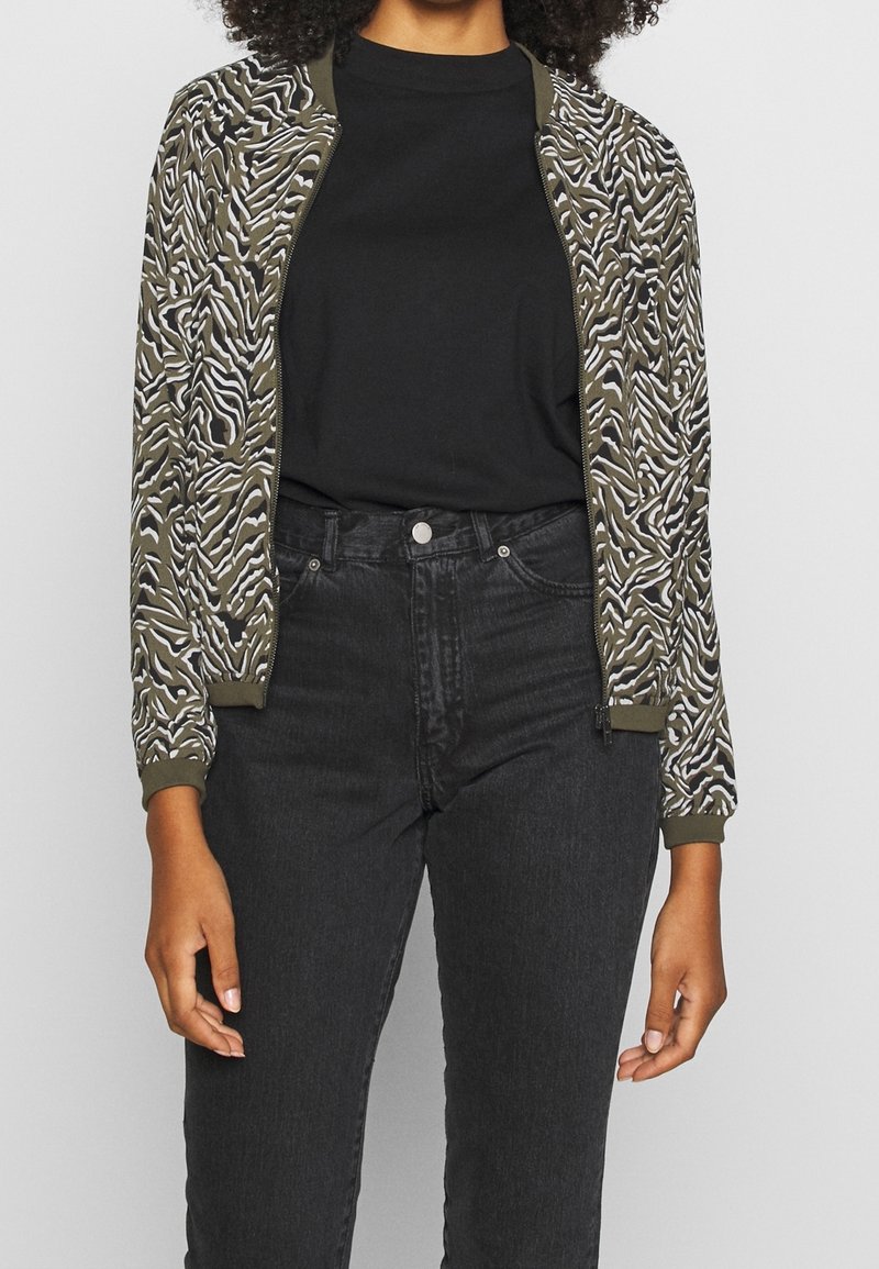 Personne portant un t-shirt noir, un jean noir taille haute et une veste ouverte verte et blanche à motif zèbre.