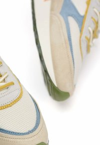 Zapatillas con parte superior de malla en crema con acentos en azul y amarillo, detalles en ante y una suela de goma verde. Líneas de diseño distintivas.