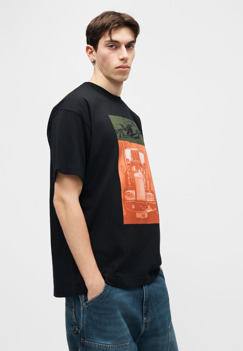 Giovane con capelli castani corti che indossa una maglietta nera oversize con grafica di camion arancione e verde, jeans blu, in piedi davanti a uno sfondo bianco semplice.