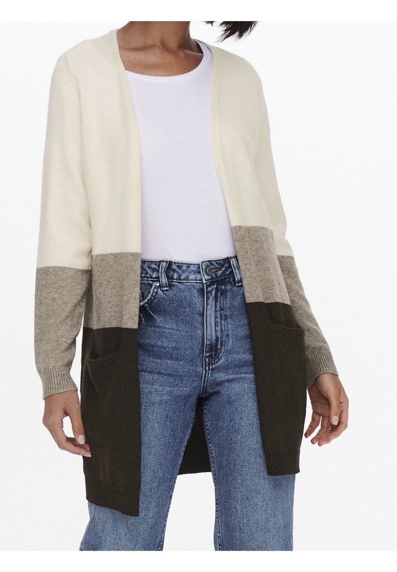 Femme portant un long cardigan avec blocs de couleurs blanc, beige et brun foncé sur une chemise blanche et un jean bleu.