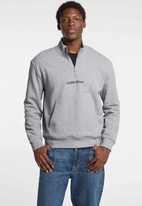 Pelēks zip-up džemperis ar augstu apkakli, ar priekšējo kabatu un melnu "GUESS JEANS" logotipu uz krūtīm, valkāts ar ziliem džinsiem.