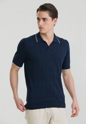 Mann trägt ein marineblaues, kurzärmeliges, gestricktes Polo mit vertikalen Streifen und weißem Rand, schaut seitlich und steht vor einem einfarbigen hellgrauen Hintergrund.