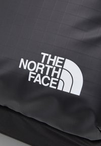 Svart ryggsäck med en strukturerad yta och vitt logotyp. Designen inkluderar ett subtilt rutmönster och en hållbar svart tygbas.