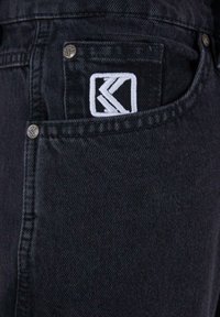 Jeans in denim nero con tasca laterale e logo ricamato bianco. Il tessuto ha una texture liscia e una vestibilità classica.
