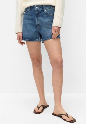 Osoba ubrana w niebieskie krótkie spodenki dżinsowe, kremową bluzkę z długim rękawem i brązowe japonki, stojąca na jednolitym białym tle.