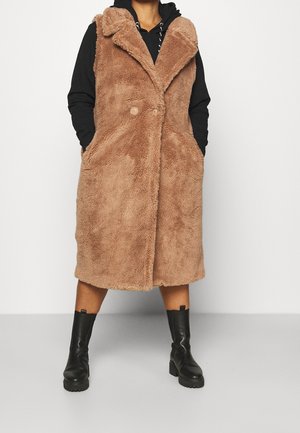 Femme portant un long manteau teddy marron, boutonné et sans manches, par-dessus un sweat à capuche noir et des bottines noires, debout devant un fond uni.