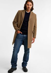 Brązowy płaszcz o dopasowanym kroju, noszony na granatowym swetrze, w połączeniu z niebieskimi dżinsami i czarnymi butami. Gładka tekstura i klasyczny design.