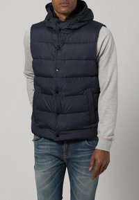 Mann trägt eine dunkelblaue Steppweste mit Kapuze über einem hellgrauen Langarmshirt und blauen Jeans, steht mit einer Hand in der Tasche.