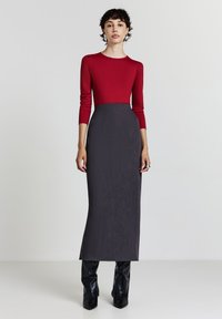 Donna in piedi che indossa un top rosso attillato a maniche lunghe con una gonna midi grigio scuro a vita alta e stivali neri con tacco.