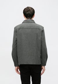 Jachetă textilă gri cu model houndstooth, mâneci cu nasturi, șliț spate și guler. Croială simplă, adaptată, fără închideri vizibile.