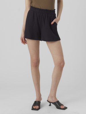 Donna che indossa pantaloncini neri larghi con vita elastica e mule nere aperte sulle punte con tacco, in piedi con una mano in tasca su sfondo semplice.