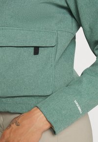 Mintgrön jacka med ett texturerat material, som har en framficka och en synlig "WINDWALL"-etikett på ärmen.