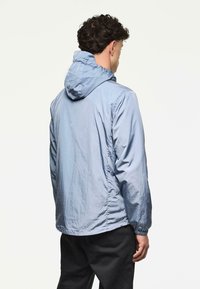 Man met een lichtblauwe gewatteerde windjack en zwarte broek, gezien vanaf de achterkant tegen een eenvoudige lichte achtergrond.