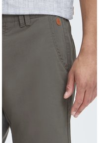Gray cotton pants with a smooth texture, featuring a side pocket and a brown tag accent near the waistband. Tailored fit.
