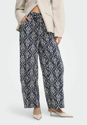 Femme portant un pantalon large bleu marine à motif floral crème, un blazer beige et des talons beiges pointus, les mains dans les poches.