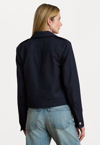 Donna con capelli biondi che indossa una giacca blu scuro e jeans a vita alta di un blu chiaro, vista di spalle su uno sfondo bianco semplice.