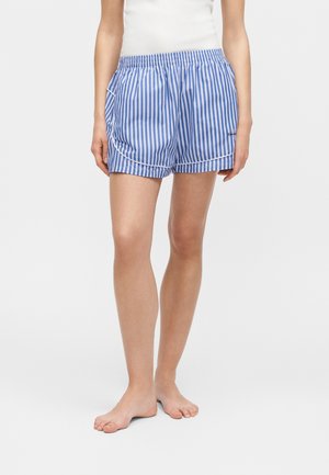 Personne portant un short à taille élastique rayé verticalement bleu et blanc, debout pieds nus sur un fond clair uni.