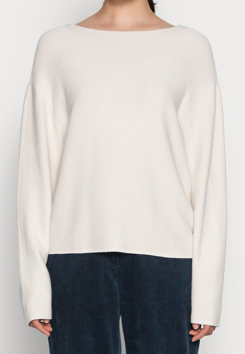Eine Frau trägt einen lockeren, langärmeligen, cremefarbenen Strickpullover, kombiniert mit dunkelblauen Cordhosen, und steht vor einem schlichten Hintergrund.