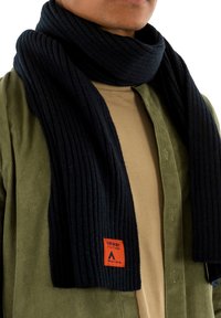 Écharpe en tricot côtelé marine avec une étiquette orange portant la mention "SUPERDRY". Portée sur une chemise beige et une veste en velours côtelé verte. Texture douce.