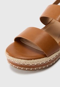 Barna bőr szandál, amely két széles pánttal és fonott juta espadrille talppal rendelkezik. Simább textúrájú, platform kialakítással.