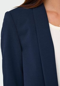 Naval blazer med en glat tekstur og strukturerede reverser, har et enkelt knapluk, parret med en cremefarvet top under.