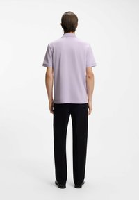 Liliowa koszulka polo o prostym kroju i krótkich rękawach, połączona z czarnymi spodniami, widziana od tyłu na jednolitym tle.